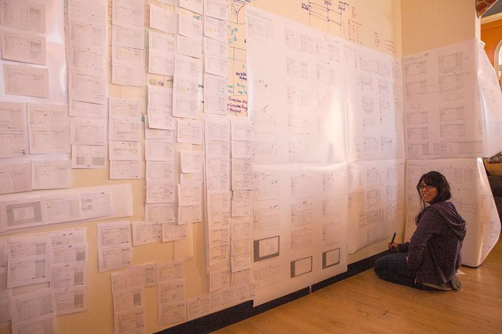 A smiling woman kneels infront of a wall filled with large scale design print-outs